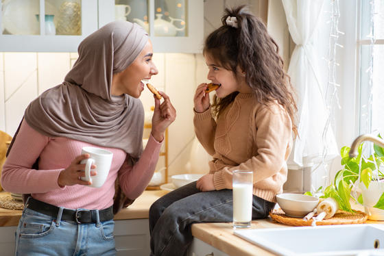 Muslimische Mutter mit Hijab und ihre kleine Tochter essen Snacks in der Küche.